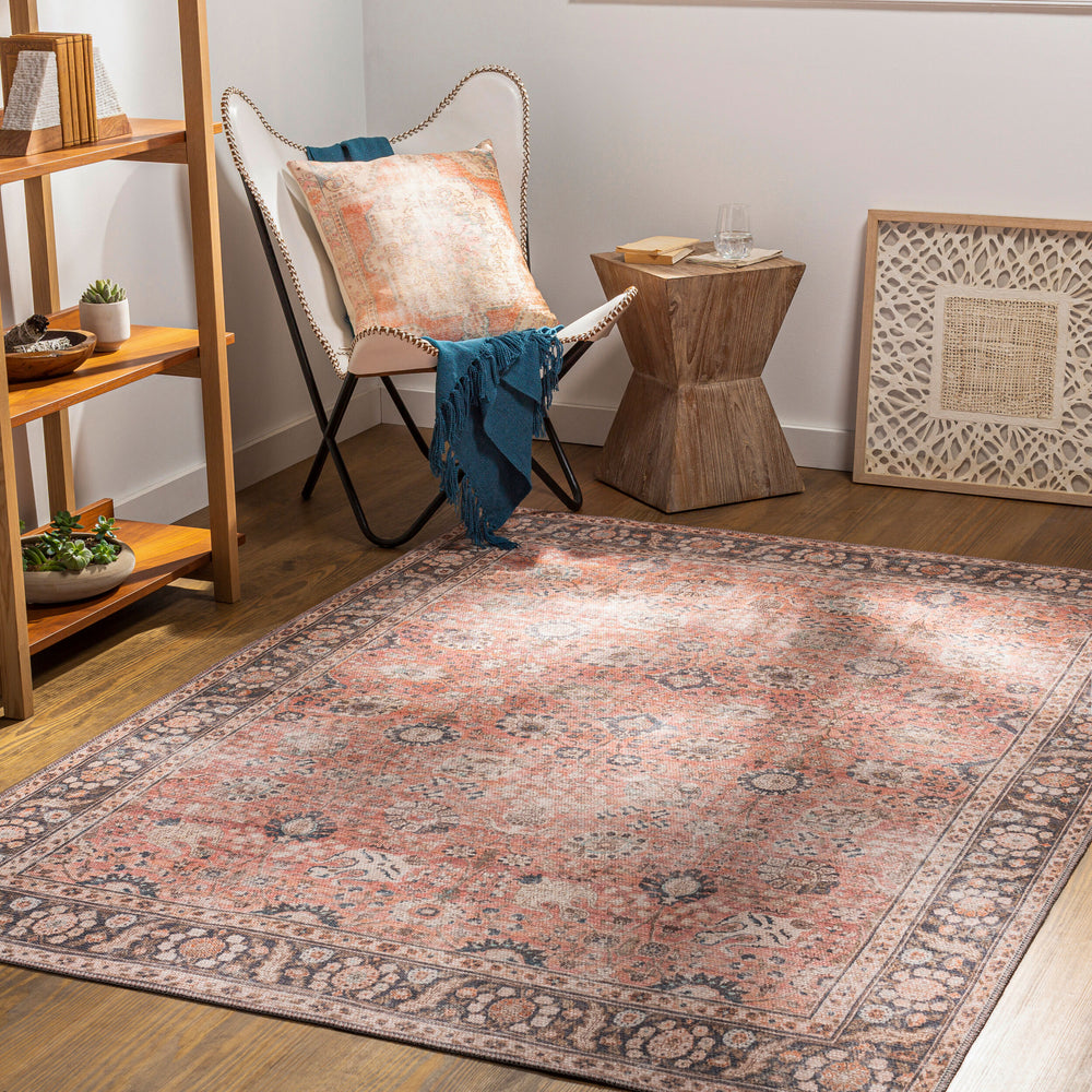 A traditional patterned rug in dusty coral, rust, medium brown, dark brown, tan, light beige, blue, charcoal, and black colors displayed in a room setting with a chair and side table.